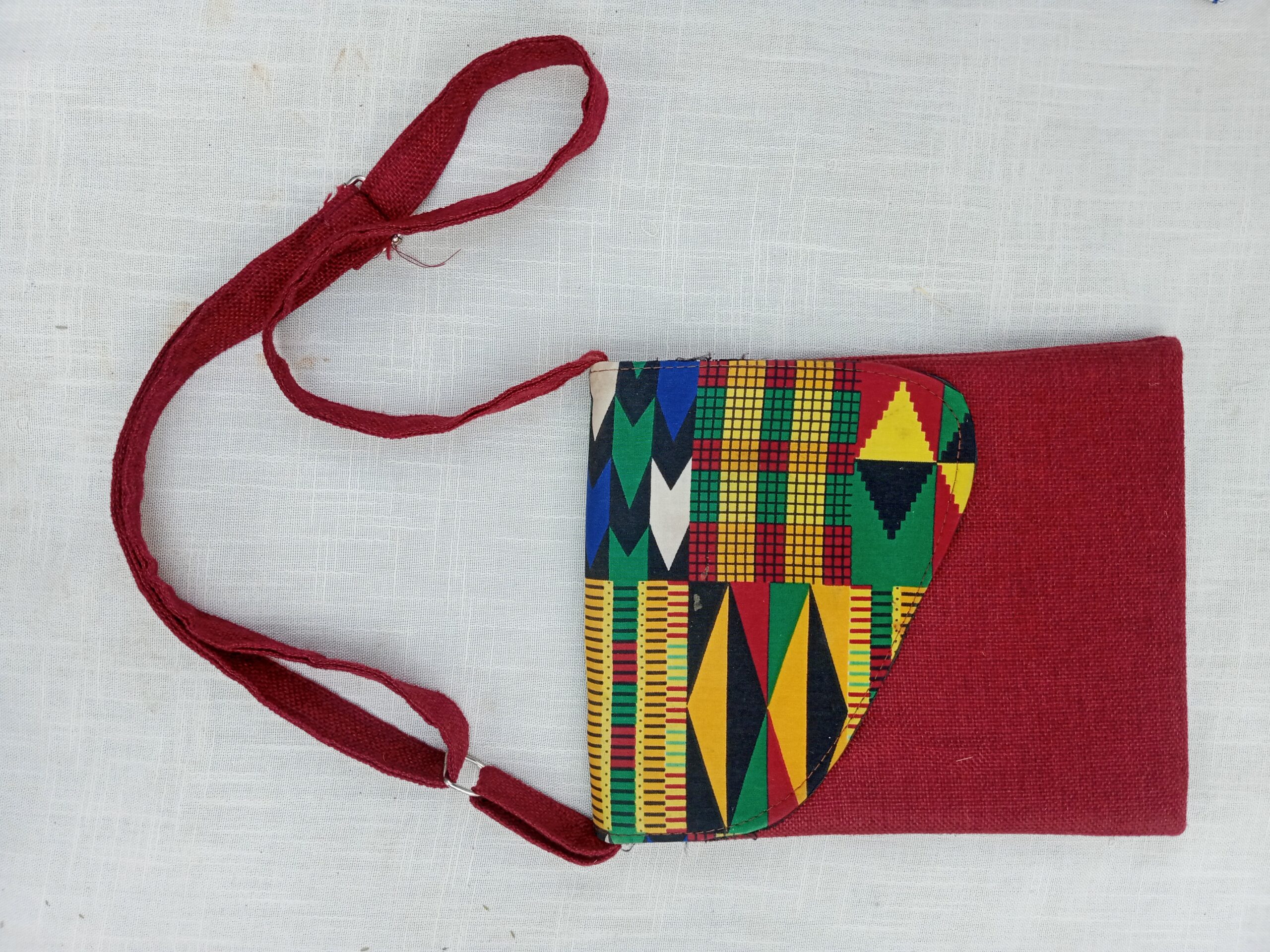 A compact crossbody bag featuring a bold African wax-print (Ankara/Kente-style) flap set against a rich burgundy woven fabric body, finished with a matching adjustable shoulder strap — a vibrant, everyday statement piece.