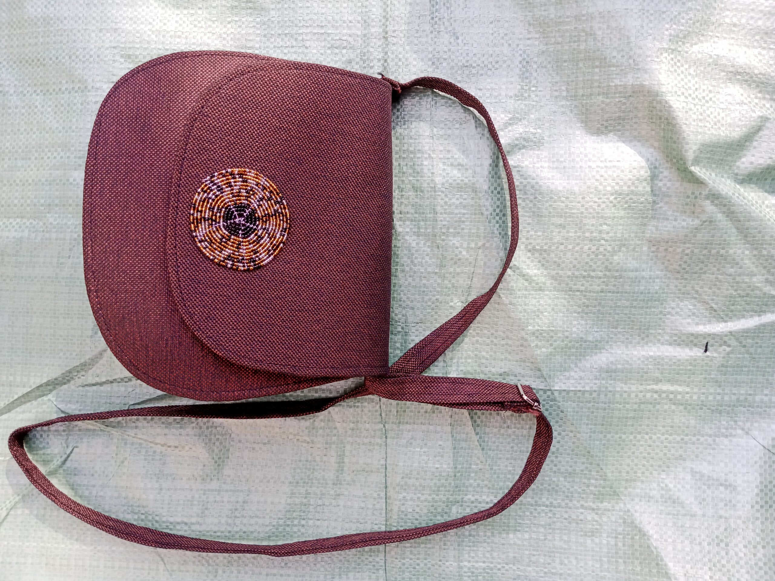 A compact crossbody bag featuring a bold African wax-print (Ankara/Kente-style) flap set against a rich burgundy woven fabric body, finished with a matching adjustable shoulder strap — a vibrant, everyday statement piece.